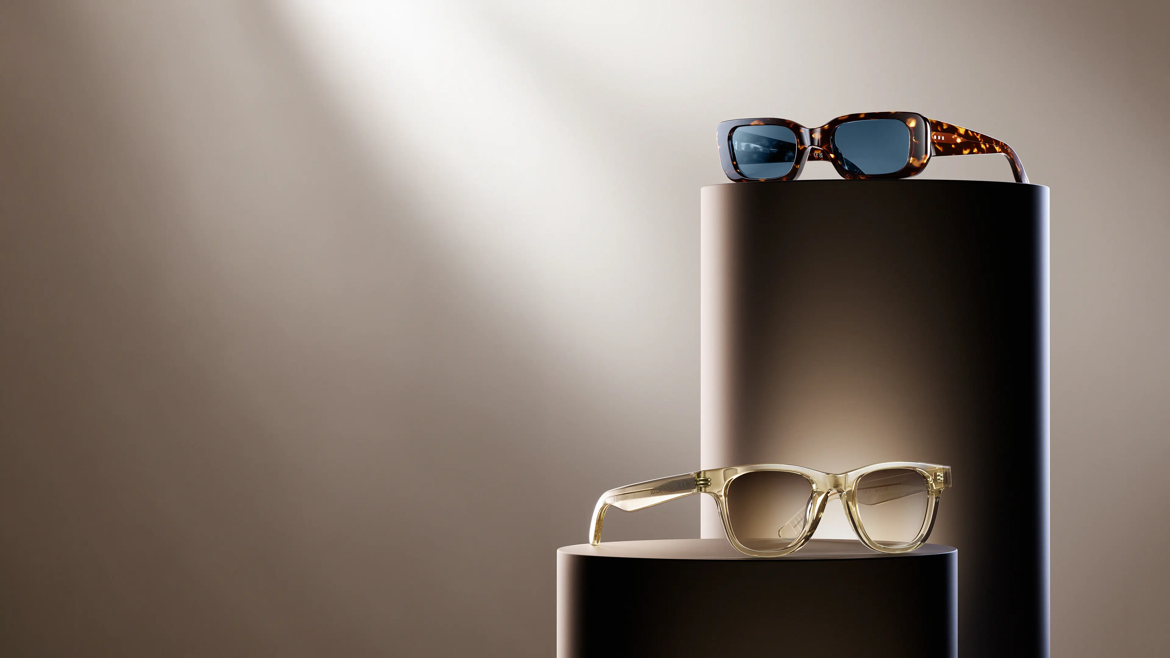 Tortoiseshell and clear acetate sunglasses displayed on cylindrical pedestals with warm studio lighting